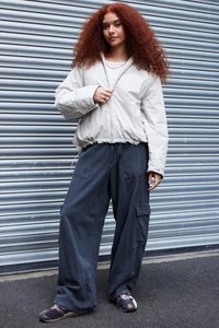 Giacca bomber grigia chiara, pantaloni cargo scuri e larghi, e scarpe nere multitonali. Capelli rossi ricci, gioielli minimal. Lo sfondo presenta un muro in metallo ondulato.