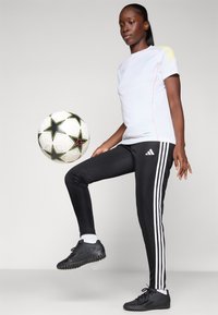White athletic shirt with colored accents and short sleeves, paired with black pants featuring white stripes. Black soccer ball nearby.