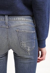 Bledě modré džíny z denimu mají malou poškozenou oblast na zadní kapse a standardní švy. Tkanina je měkká s mírným elastickým strečem.