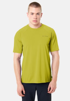 Giovane uomo con capelli corti che indossa una maglietta sportiva verde lime a maniche corte e pantaloni scuri, in piedi contro uno sfondo chiaro uniforme.