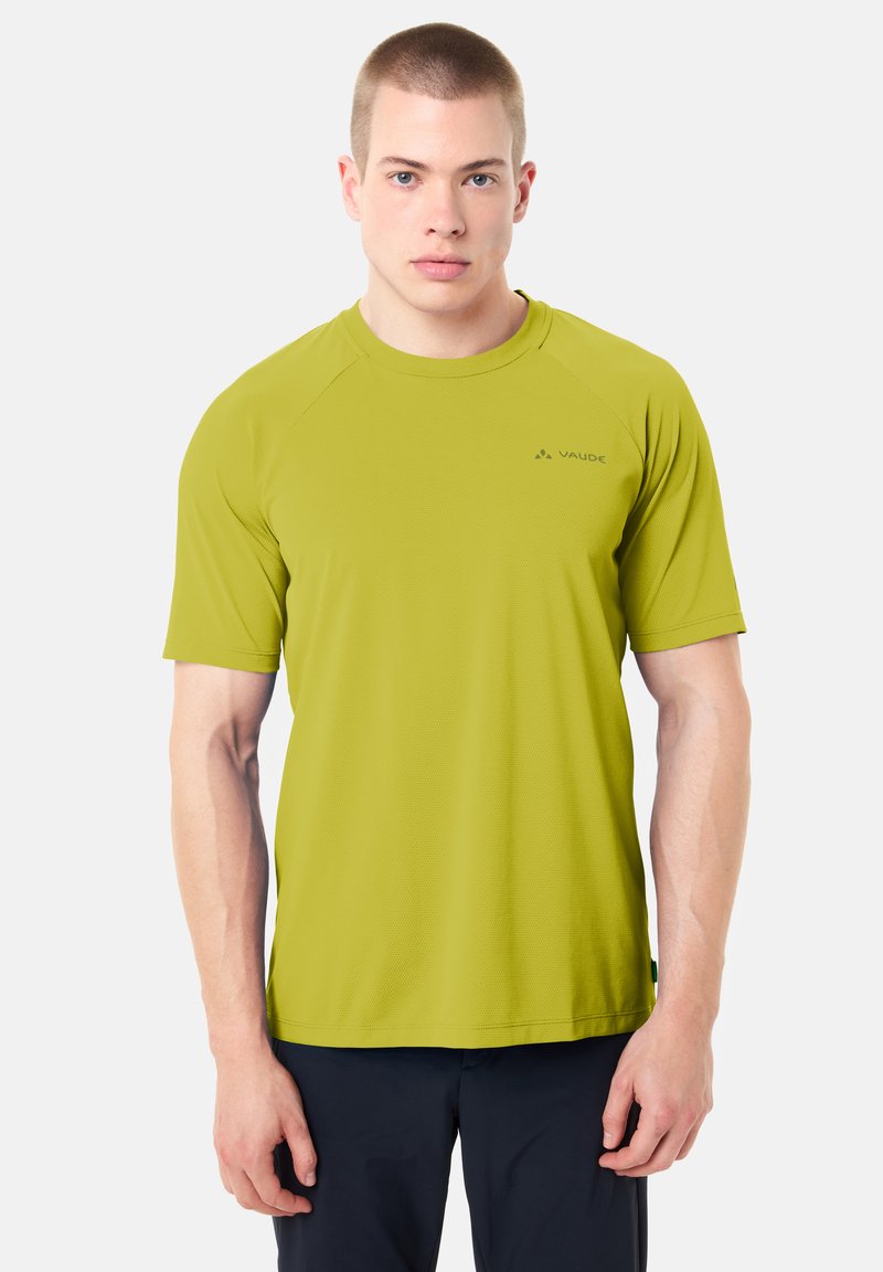 Jeune homme aux cheveux courts portant un t-shirt de sport vert citron à manches courtes et un pantalon foncé, debout devant un fond clair uni.