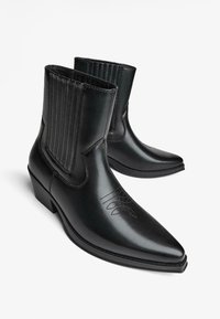 Črne usnjene gležnjarje s koničastim nosom, rebrastimi elastičnimi paneli in okrasnim šivanjem. Nizka nabirana peta in gladka površina.