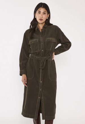 Donna che indossa un lungo vestito di velluto a coste verde oliva scuro con tasche frontali, bottoni al centro, cintura in vita e maniche lunghe.