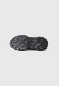 Svart sneakersåle med bølgede, teksturerte mønstre og "GUESS"-logo preget nær hælen, fotografert på en hvit bakgrunn.
