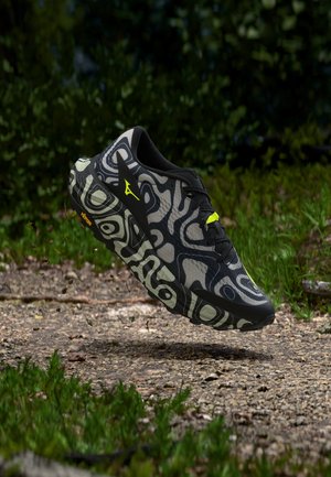 Scarpa da corsa nera e grigia con motivo e dettagli giallo neon che fluttua sopra un sentiero di terra circondato da fogliame verde.