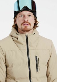 Beige isolierte Jacke mit hohem Kragen und Reißverschluss, ausgestattet mit einer linken Brusttasche und schwarzen Akzenten, getragen mit einer Skibrille und einer Beanie.