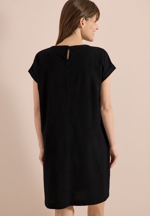 Kobieta o prostych brązowych włosach, ubrana w czarną teksturowaną sukienkę z krótkim rękawem, lekko odwrócona na bok na beżowym tle.