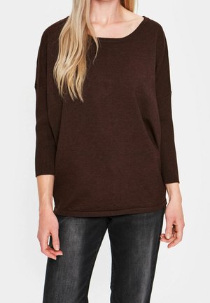 Femme portant un pull ample à manches longues marron foncé avec des manches côtelées et un jean noir délavé, debout sur fond blanc.