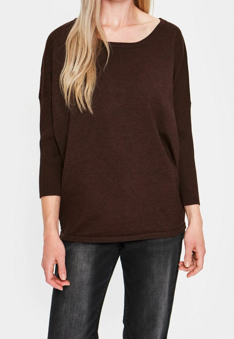 Femme portant un pull ample à manches longues marron foncé avec des manches côtelées et un jean noir délavé, debout sur fond blanc.
