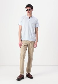 Svetlomodrá polo tričko s krátkymi rukávmi, s malým logom na hrudi. Béžové nohavice a hnedé topánky. Model nosí tmavé slnečné okuliare.
