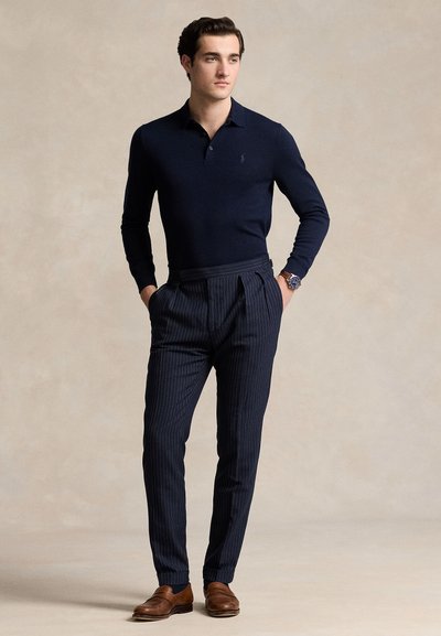 Hombre que lleva un polo azul marino de manga larga, pantalones azul marino con rayas finas, zapatos de cuero marrón y reloj de pulsera, de pie con las manos en los bolsillos.