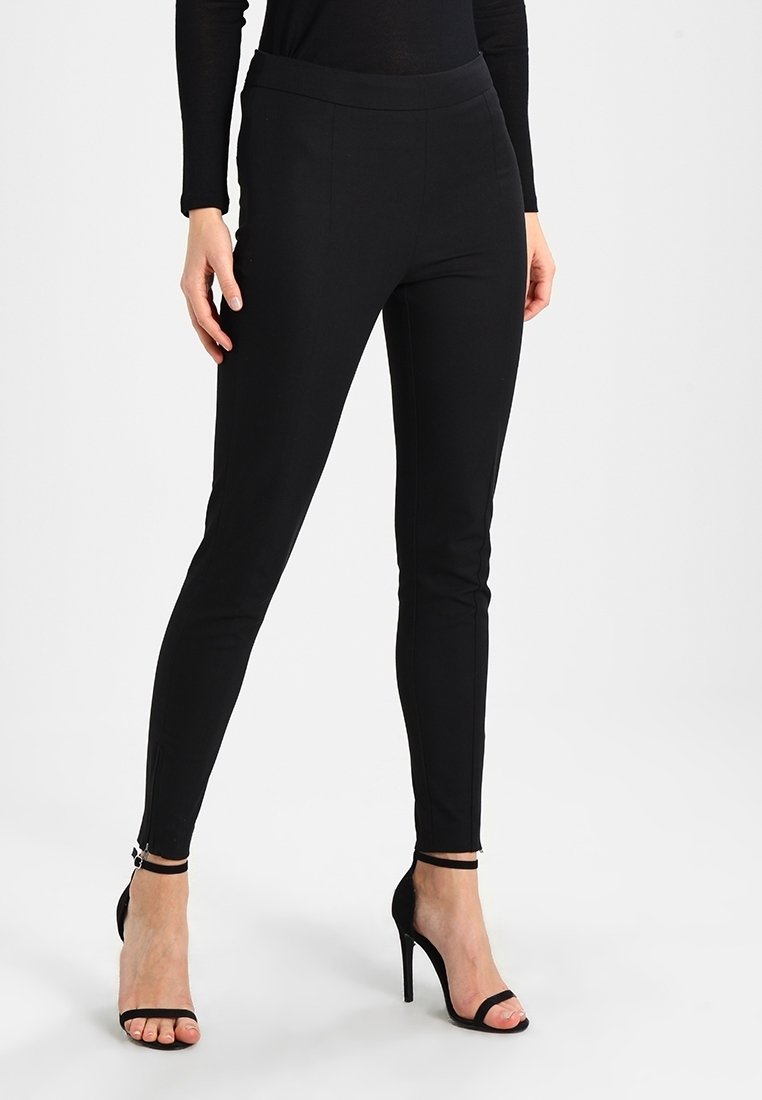 Femme portant un pantalon noir ajusté et des sandales noires à talons hauts avec des brides à la cheville, debout devant un fond uni.