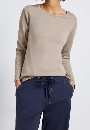 Kobieta w jasno beżowej bluzce z długim rękawem i granatowych spodniach z sznurkiem, stojąca z rękami w kieszeniach na białym tle.