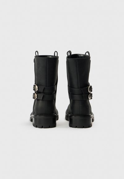 Černé kožené kotníkové boty se dvěma bočními pásky, kovovými ozdobami a robustní podrážkou. Hladká textura s kulatou špičkou.