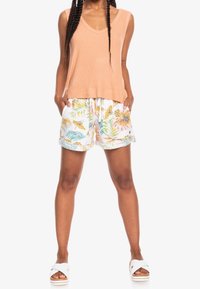 Débardeur en maille pêche avec col en V, associé à un short blanc orné de motifs colorés de feuilles tropicales et de fleurs. Sandales blanches à enfiler.