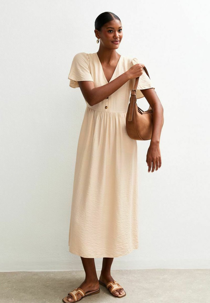 Beige midi-jurk met korte, geplooide mouwen, v-hals en knopen op het lijf. Gecombineerd met een bruine schoudertas en sandalen. Soepele textuur.