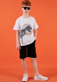 Garçon aux cheveux roux portant des lunettes de soleil noires, un t-shirt blanc avec une imprimé de squelette de T-Rex, un short noir et des baskets blanches, debout sur un fond orange.