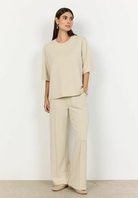Beige oversized t-shirt and wide-leg pants; smooth fabric with subtle drape; pants feature front seams; model stands with one hand in pocket.
