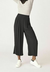 Široké, skladané kalhoty z tmavě šedé látky, s vysokým pasem a hladkou texturou, doplněné bílými teniskami.