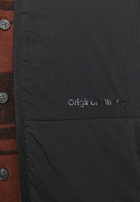 Zwarte jas met een gestructureerde afwerking, voorzien van een opvallende rits, met een bruin en zwart geruit patroon zichtbaar eronder, en het 'Originals Studio' logo.