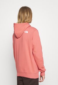 Osoba s dlouhými blond vlasy, oblečená v korálově růžové mikině The North Face, otočená zády k jednobarevnému bílému pozadí.