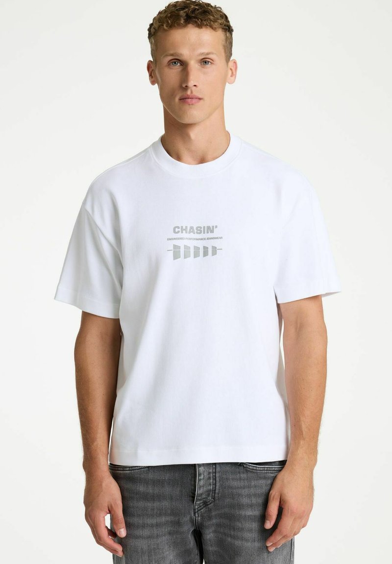 Camiseta blanca de algodón con cuello redondo, mangas cortas y texto gráfico reflectante en gris. Combinada con pantalones vaqueros gris oscuro con textura.