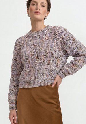 Vrouw draagt een meerkleurige lavendel gebreide trui gecombineerd met een bruine rok, staand met één hand op haar heup, lichte achtergrond.