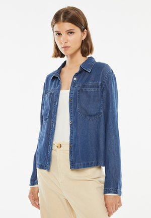 Camicia in denim di colore blu scuro con maniche lunghe, chiusura a bottoni davanti, due tasche sul petto e colletto a punta. Tessuto liscio, vestibilità casual.