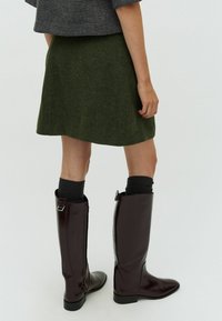 Stivali marroni lucidi fino al ginocchio con una silhouette elegante, dotati di una cerniera laterale e un piccolo dettaglio in metallo a forma di fibbia, abbinati a una gonna verde con texture.