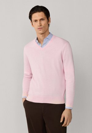 Man wearing light pink V-neck sweater over light blue dress shirt and dark brown pants, standing with one hand in pocket against plain background.