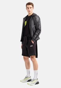 Giacca nera lucida, t-shirt nera con logo verde, pantaloni corti neri, calze bianche e sneakers grigio-neri con accenti neon.