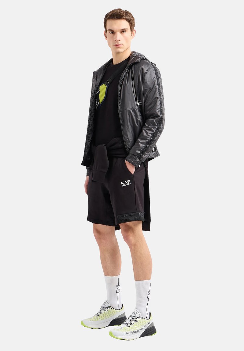 Giacca nera lucida, t-shirt nera con logo verde, pantaloni corti neri, calze bianche e sneakers grigio-neri con accenti neon.