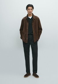 Bruine suede jas over een zwarte polo, gecombineerd met donkere broeken en bruine schoenen. De jas heeft een knoopsluiting aan de voorkant en een ontspannen pasvorm.