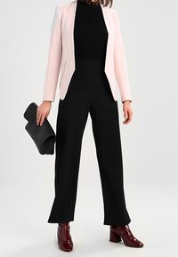 Bleknad rosa kavaj med vita detaljer, svart ribbat linne, svarta vida byxor och bordeauxfärgade klackstövlar; håller en svart clutch.