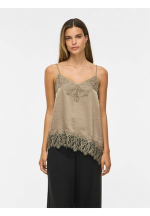 Frau trägt eine taupefarbene Satin-Camisole mit Spitzenbesatz und dünnen Trägern sowie schwarze weit geschnittene Hose vor einem schlichten weißen Hintergrund.