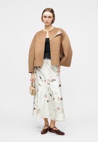Frau trägt eine übergroße Kamelhaarjacke mit einer Brosche, ein schwarzes Oberteil, einen floralen Midirock, eine beige Handtasche und braune Mary Jane Schuhe.
