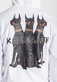 Biely mikina s veľkým grafickým zobrazením troch dobermanov so zlatými goliermi a textom "KAVANAGH" v čiernom geometrickom písmene nižšie.