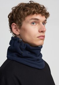 Giovane con capelli ricci che indossa un collo in pile blu navy con cordino regolabile, vestito con una maglietta nera, che guarda a destra.