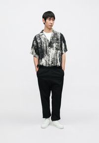 Kurzärmliges Hemd mit schwarz-weißem Muster, schwarze Jogginghose und weiße Sneaker. Das Hemd zeigt ein abstraktes Design; die Hose hat seitliche Streifen.