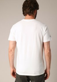 T-shirt en coton blanc avec un col rond et des manches courtes. Présente un petit détail textuel sur la manche droite. Texture lisse, coupe décontractée.