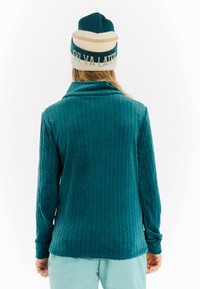 Persoon in een turquoise textuur jack en een turquoise-creme muts met de tekst "Ski Ya Later", staande met de rug naar de camera tegen een witte achtergrond.