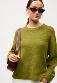 Ženska nosi olivnozeleni pleteni pulover, crne mačje sunčane naočale, naušnice na obruče i nosi tamnocrvenu kožnu torbu preko ramena.