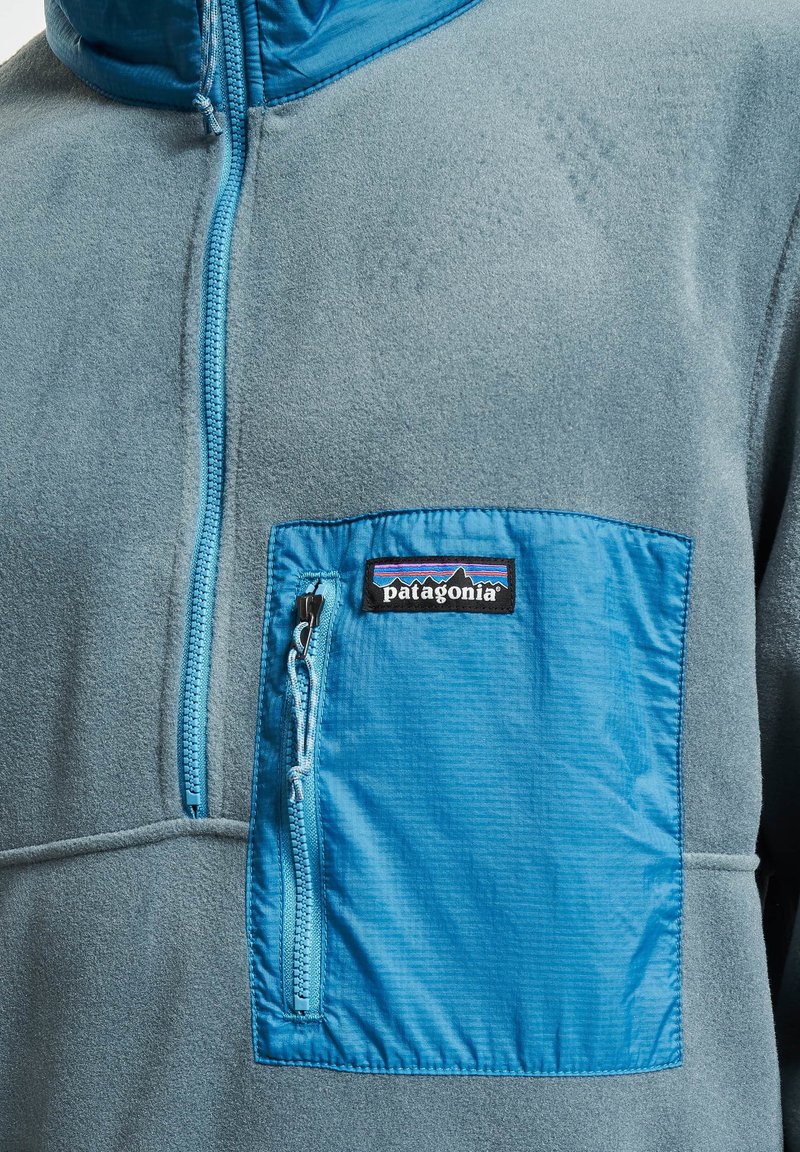 Pullover in pile di colore azzurro chiaro con tasca con zip di colore blu scuro a contrasto. Presenta una texture liscia e il logo Patagonia in evidenza.