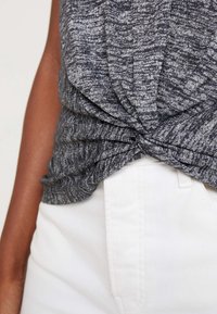 Primo piano di un top a maglia grigio mélange con un dettaglio a twisted front indossato con pantaloni bianchi a vita alta.