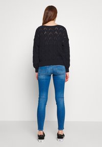 Suéter negro de punto con un patrón de diamante texturizado, combinado con jeans ajustados azules y zapatillas negras. El suéter tiene un corte relajado y puños acanalados.