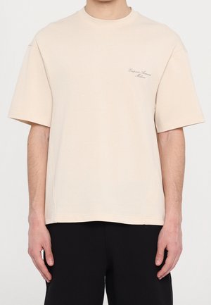 Homme portant un t-shirt beige oversize avec le logo "Emporio Armani Milano" sur la poitrine gauche et un pantalon noir, debout devant un fond uni.