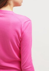 Ung person som står med sidan mot kameran och bär en ljusrosa långärmad skjorta mot en enfärgad ljus bakgrund.