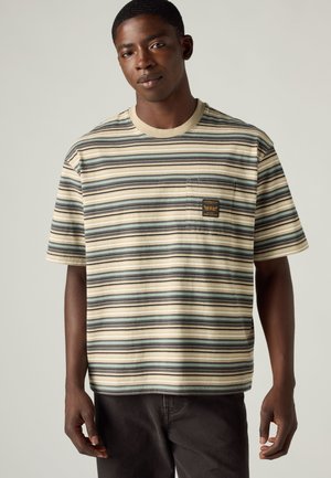 Joven con camisa de manga corta a rayas beige, azul y negra con bolsillo en el pecho y pantalones oscuros sobre un fondo claro y liso.