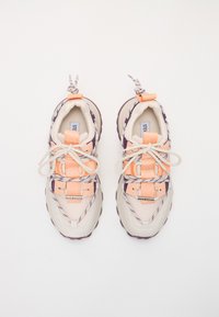 Sneaker con tomaia in mesh beige, accenti arancioni e suola in gomma viola. Presenta un sistema di lacci unico e un marchio sulla linguetta.