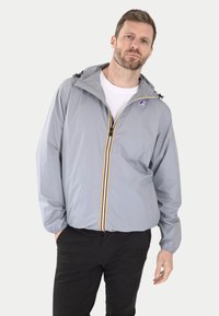 Chaqueta impermeable gris claro con capucha ajustable, frente con cremallera y rayas de acento amarillas. Cuenta con puños elásticos y un parche con logo.